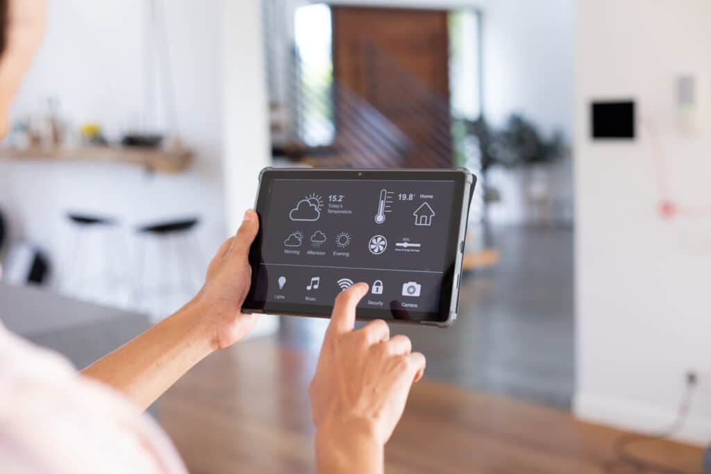 Personne tenant une tablette avec une interface de maison connectée. L'écran affiche météo (15.2°C), température intérieure (19.8°C), économie d'énergie (40%) et icônes pour lumières, musique, sécurité, caméra, et Wifi sur lequel un doigt appuie. Arrière-plan flou d'un intérieur moderne.