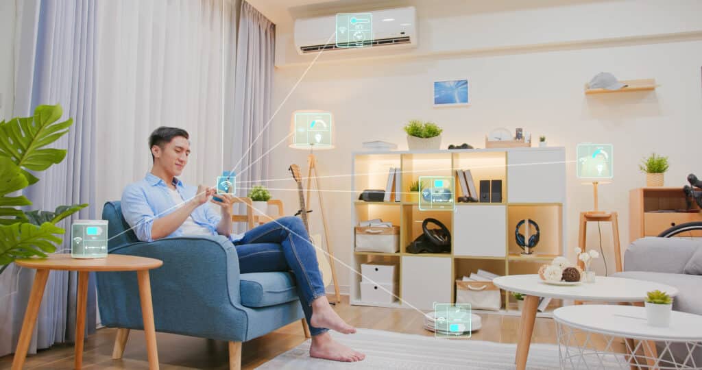 Homme souriant sur un fauteuil, contrôlant sa maison intelligente via smartphone; icônes AR d'objets connectés (clim, lampes, robot aspirateur) apparaissent.