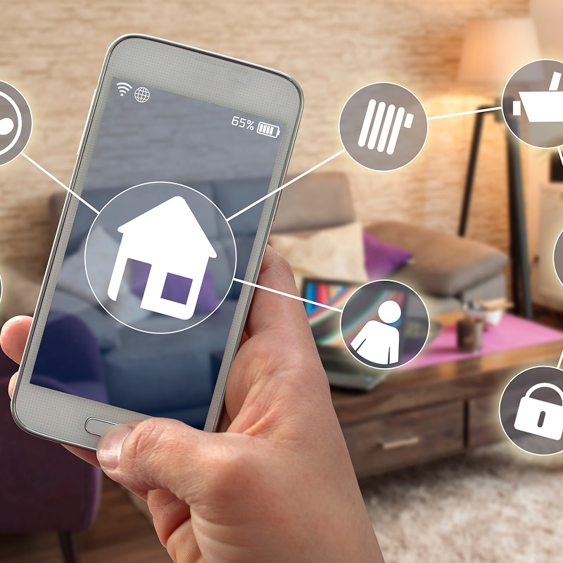 Main contrôlant une application domotique sur smartphone (batterie 65%). Écran centralise une maison et icônes : chauffage, sécurité, présence.