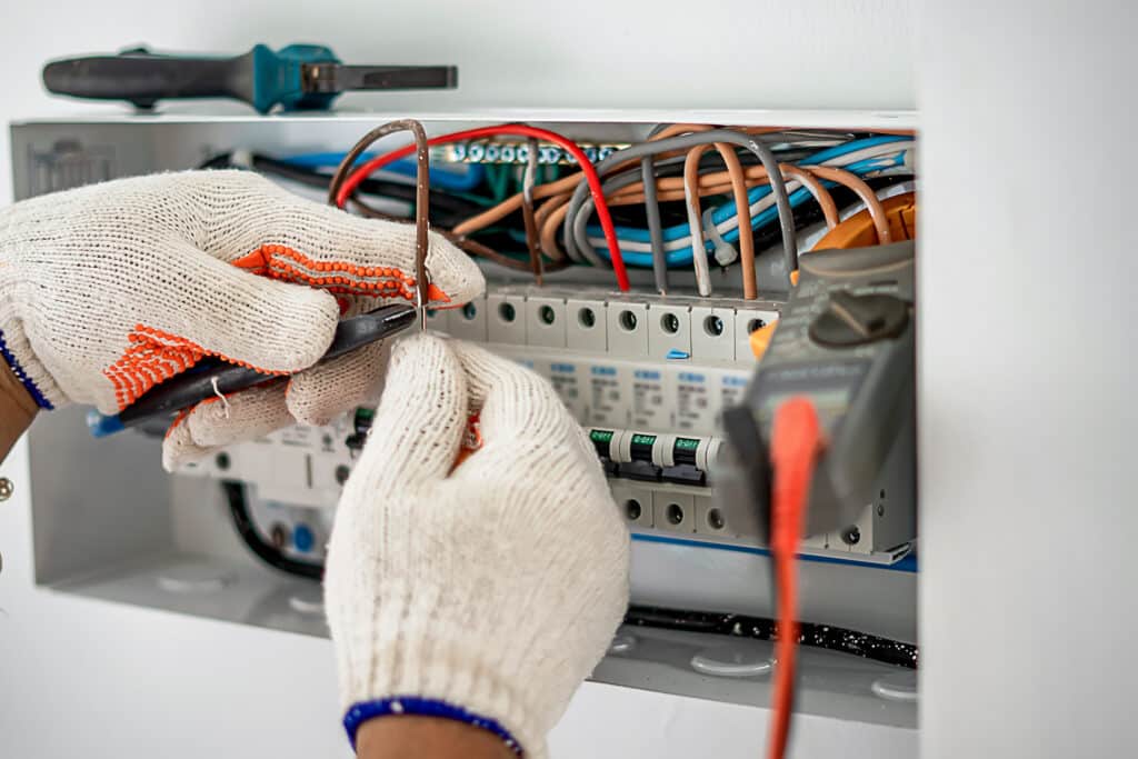 Électricien en gants de travail connectant des fils colorés dans un tableau électrique. Disjoncteurs affichent "O-OFF". Multimètre visible.
