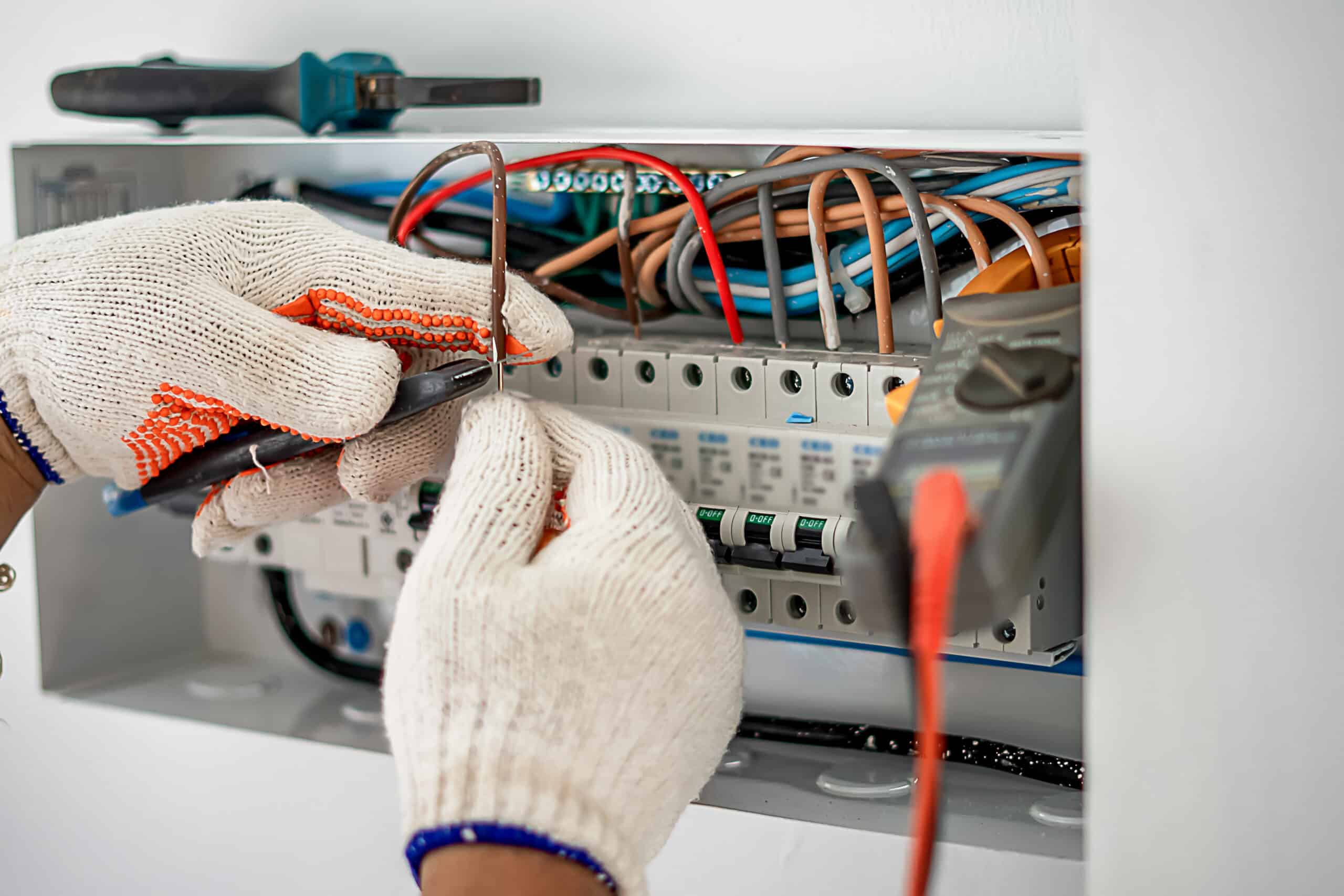 Électricien en gants de travail connectant des fils colorés dans un tableau électrique. Disjoncteurs affichent "O-OFF". Multimètre visible.