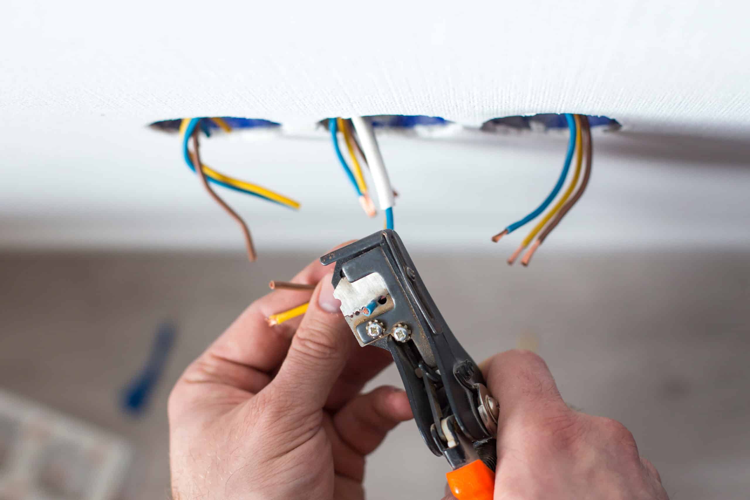 Électricien dénudant des fils électriques (bleu, jaune, marron) avec une pince, pour installer des prises ou luminaires dans un mur blanc.