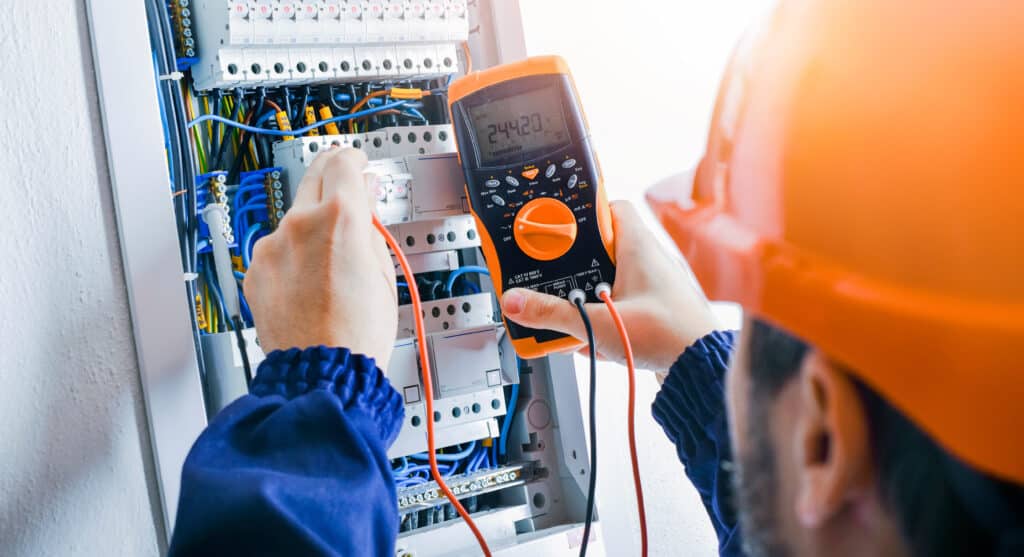 Électricien mesurant la tension (244.20 V) d'un tableau électrique avec disjoncteurs "B 16" et câblage. Sécurité chantier.