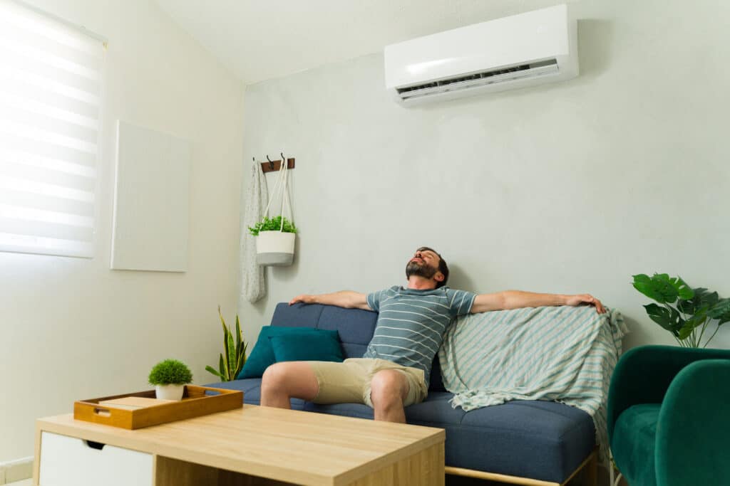 Homme barbu détendu sur un canapé bleu dans un salon moderne. La climatisation affiche 24°C, apportant fraîcheur. Lumière naturelle, plantes.