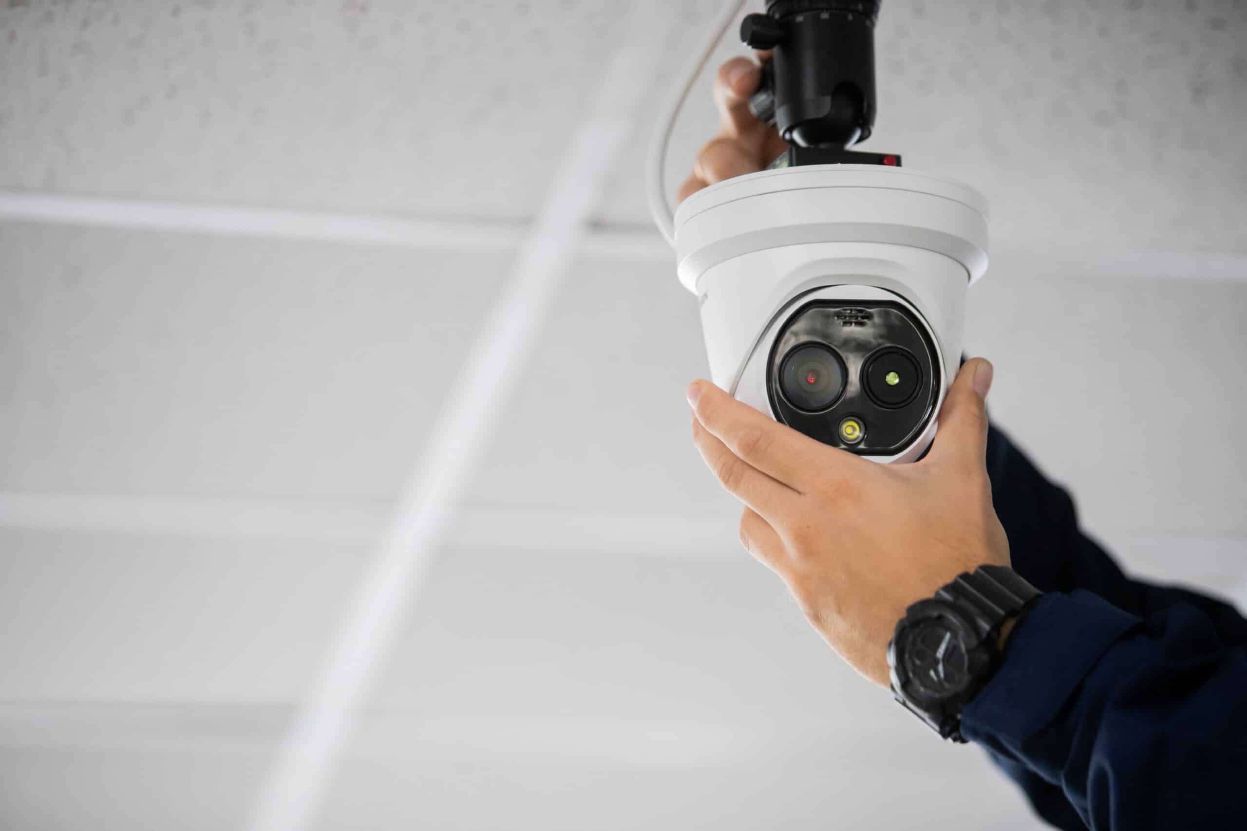 Mains d'un technicien ajustant une caméra de surveillance blanche fixée au plafond, avec des voyants rouge et vert allumés.