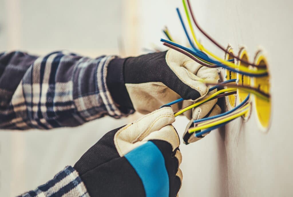 Mains électriques: fils colorés (jaune, bleu, marron, vert) installés dans un mur par des mains gantées. Installation électrique.
