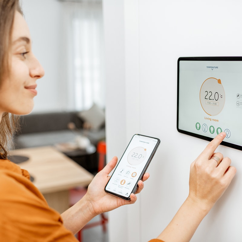Femme souriante contrôlant la température (22.0°C) du salon sur une tablette murale et un smartphone. Interface domotique.