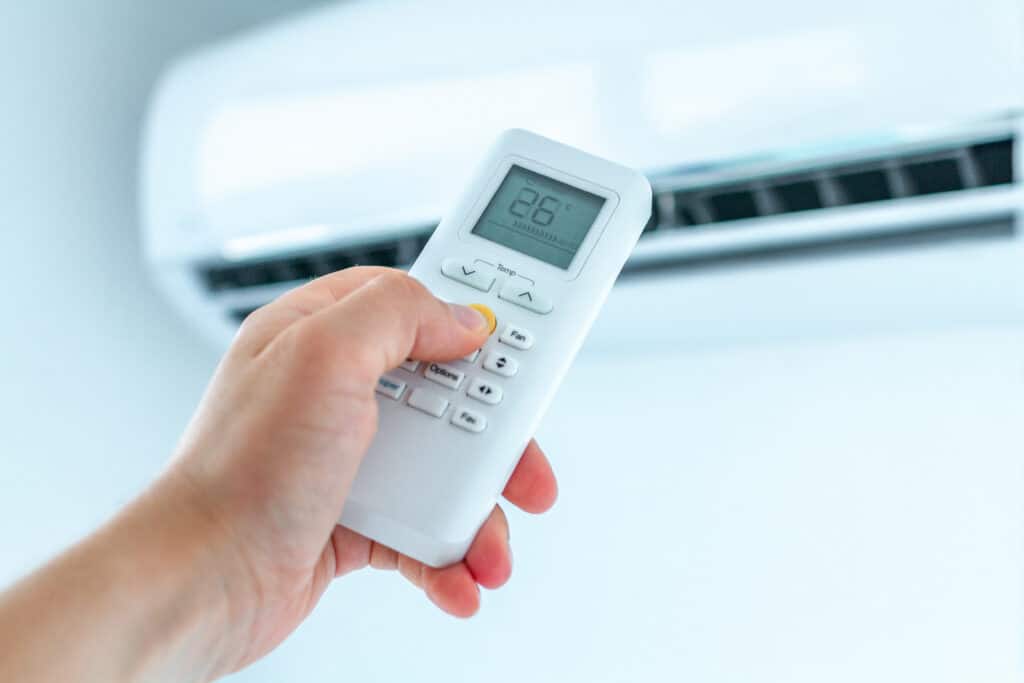 Main pressant la télécommande d'un climatiseur affichant 26°C AUTO et un ventilateur. Le climatiseur mural blanc est visible en arrière-plan, ajustant la température ambiante.